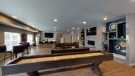 This single-floor amenity space is thoughtfully designed as a multifunctional gathering hub, blending modern aesthetics with a welcoming, social atmosphere. The open-plan layout flows seamlessly across distinct activity zones, all unified by wood-look flooring and recessed lighting throughout.

The central lounge area anchors the space with a generously sized dark leather sectional sofa oriented around a tiled fireplace feature wall. Built-in shelving with bold blue accent panels flanks the fireplace, adding visual interest and display space. A wall-mounted flat-screen TV above the fireplace ensures this area functions as both a relaxation and entertainment focal point.

Adjacent to the lounge, a full-length shuffleboard table serves as the centerpiece of the game zone, accompanied by additional seating. A separate pub-style nook features high-top tables with bar stools, wall-mounted artwork, and a dartboard — creating an ideal casual games corner with natural light from nearby windows.

The kitchenette is well-appointed with light wood cabinetry, a white countertop island with bar seating, a stainless steel refrigerator, a dishwasher, a water dispenser, and a mounted television — making it well-suited for hosting and catered events. The overall design palette of neutral grays, warm wood tones, and rich blue accents creates a cohesive, contemporary ambiance throughout.