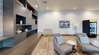 Located at 1700 Newbury Park Dr in San Jose, California, this single-floor community space offers a generously sized, open-plan layout that seamlessly integrates a fully equipped kitchen and an expansive living area. The design is distinctly contemporary, with large-format wood-look tile flooring, recessed lighting, and crisp white walls that create a bright, welcoming atmosphere throughout.

The kitchen is a standout feature, anchored by a wide island with a white quartz countertop and seating for multiple guests at warm wood barstools. Stainless steel appliances, white cabinetry, and a decorative patterned tile backsplash complete the functional yet stylish cooking area. A bold gold pendant light adds a striking accent overhead.

The living area is thoughtfully zoned into multiple lounge and social spaces. A built-in shelving wall with a large flat-screen TV serves as the focal point of the media zone, flanked by patterned wallpaper for added visual interest. Comfortable seating arrangements, including plush sofas and accent chairs, invite relaxation, while whimsical hanging rattan swing chairs near floor-to-ceiling windows add a playful, distinctive touch. Natural light pours in through large windows, offering views of mature trees and the surrounding neighborhood. A dedicated lounge nook with built-in shelving and curated décor completes the space.