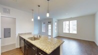 This single-floor space showcases a thoughtfully designed open-concept layout that seamlessly connects the kitchen and living areas. The kitchen serves as the centerpiece, featuring a large island adorned with striking granite countertops in warm cream and gold tones, complemented by rich dark-espresso cabinetry with sleek hardware. A stainless steel sink sits within the island, and three elegant pendant lights hang overhead, providing both task lighting and a stylish focal point. Recessed lighting throughout adds to the bright, welcoming atmosphere. The open living area extends naturally from the kitchen, finished with warm hardwood-style flooring that flows throughout the space. Large windows along two walls flood the interior with natural light while offering views of the surrounding exterior. A glazed door provides direct access to an outdoor area, enhancing the sense of connectivity between indoor and outdoor living. The clean white walls and contemporary finishes lend the space a crisp, modern aesthetic. The overall ambiance is light, airy, and functional, making it an inviting environment well-suited for both everyday living and entertaining.