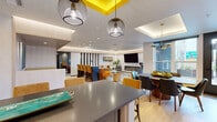 Located at 224 N Barstow St in Eau Claire, Wisconsin, this thoughtfully designed communal space offers a seamless blend of social and functional areas within a single open-concept floor. The kitchen is outfitted with stainless steel appliances, light wood cabinetry, a striking teal subway tile backsplash, dark countertops, and a large island with bar seating — all illuminated by a series of globe pendant lights suspended beneath a bold yellow ceiling accent. Adjacent to the kitchen, a spacious dining area features a modern table with seating for several guests, natural light flowing in through large windows, and a refined brass chandelier overhead. The open living room is anchored by a long linear fireplace, a wall-mounted flat-screen television, and cove lighting that runs along a tray ceiling, lending a warm and inviting ambiance. Comfortable seating arrangements and layered area rugs enhance the lounge-like feel. A separate billiards room, partially enclosed by slatted dividers, provides a more private recreational retreat with its own pendant lighting and textured carpet. Light wood flooring ties the main areas together, while the cohesive contemporary design makes this space ideal for gathering and entertaining.