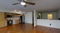 This single-floor space is designed with an open-concept layout that seamlessly connects the living area and kitchen, creating a spacious and functional environment. Warm hardwood flooring flows throughout, adding a rich, inviting character to the interior.

The kitchen is well-appointed with stainless steel appliances, including a refrigerator and dishwasher, complemented by light wood cabinetry with open shelving for easy access to everyday essentials. A mosaic tile backsplash adds a subtle decorative touch, while the countertop workspace provides ample room for meal preparation.

Overhead, a ceiling fan with an integrated light fixture anchors the main living area, promoting air circulation and ambient lighting. Track lighting installed nearby offers additional directional illumination, making the space both practical and well-lit.

A partial wall with a pass-through opening separates zones within the space while maintaining visual connectivity, enhancing the open feel of the floor plan. Crisp white walls throughout keep the atmosphere bright and neutral, offering a versatile backdrop for any interior style.

Overall, this space combines modern functionality with warm, natural finishes, resulting in a comfortable and well-designed single-floor environment suited for everyday living.