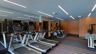 This fitness center offers a well-appointed workout environment designed for both functionality and comfort. The space is outfitted with a row of Matrix brand treadmills positioned prominently along the floor, complemented by a lineup of elliptical cross-trainers along the opposite side, providing users with a comprehensive selection of cardio equipment. Each machine features an individual display screen, allowing users to monitor their workout metrics. The ceiling is fitted with sleek linear LED light fixtures that cast even, bright illumination throughout the space, contributing to an energized atmosphere. Full-wall mirrors along one side visually expand the room and allow users to maintain proper form during exercise. The warm orange accent walls add a dynamic, motivating aesthetic contrast to the grey ceiling and dark flooring. The flooring features subtle striped detailing, adding a contemporary design element. A security camera is mounted on the ceiling, and a clearly marked emergency exit is visible, reflecting attention to safety. Overall, the space presents a clean, modern, and professionally equipped fitness facility.
