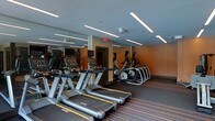 This fitness center offers a well-appointed workout environment designed for both functionality and comfort. The space is outfitted with a row of Matrix brand treadmills positioned prominently along the floor, complemented by a lineup of elliptical cross-trainers along the opposite side, providing users with a comprehensive selection of cardio equipment. Each machine features an individual display screen, allowing users to monitor their workout metrics. The ceiling is fitted with sleek linear LED light fixtures that cast even, bright illumination throughout the space, contributing to an energized atmosphere. Full-wall mirrors along one side visually expand the room and allow users to maintain proper form during exercise. The warm orange accent walls add a dynamic, motivating aesthetic contrast to the grey ceiling and dark flooring. The flooring features subtle striped detailing, adding a contemporary design element. A security camera is mounted on the ceiling, and a clearly marked emergency exit is visible, reflecting attention to safety. Overall, the space presents a clean, modern, and professionally equipped fitness facility.