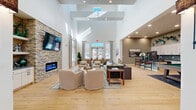 Located at 1350 W Taylor St in Sherman, Texas, this thoughtfully designed community living room offers an expansive, open-concept layout that seamlessly blends comfort and functionality. The space is defined by its dramatic soaring ceilings with skylights that flood the interior with natural light, creating an airy and welcoming atmosphere throughout.

The main seating area serves as the centerpiece, anchored by a stunning stacked-stone accent wall housing a sleek linear electric fireplace and a wall-mounted flat-screen television, flanked by built-in shelving. A sage green tufted sofa, plush swivel chairs, and a woven coffee table create an inviting conversational arrangement atop a layered area rug on warm-toned hardwood flooring that extends throughout the entire space.

Adjacent to the main lounge is a dedicated game area featuring a full-size pool table with cue rack, offering a recreational retreat within the same open floor plan. A casual dining and seating cluster near large windows provides additional gathering space bathed in natural light.

A built-in coffee and refreshment bar with stone-slab backsplash, light wood cabinetry, and illuminated open shelving adds a practical yet stylish amenity. Recessed lighting and strategically placed wall sconces complement the natural light, ensuring a warm ambiance at any hour. The cohesive neutral palette with earthy green accents ties the entire space together elegantly.