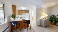 This single-floor residence offers a practical and well-organized layout designed for comfortable everyday living. The kitchen is equipped with warm medium-toned wood cabinetry providing ample upper and lower storage, white appliances including a refrigerator, electric range, dishwasher, and a single-basin sink positioned beneath a window that allows natural light to fill the space. Light wood-look vinyl flooring runs throughout the kitchen, lending a clean, modern contrast to the cabinetry.

A peninsula with a white countertop extends from the kitchen, accommodating two bar-height stools and creating a seamless transition into the living area. The living space features neutral carpeting and connects openly to the sleeping area, which is generously proportioned to accommodate a full bed and additional seating. The bedroom benefits from a calm, understated palette with soft carpet underfoot.

The bathroom includes a full-size soaking tub with an overhead shower, white tile surround, and chrome fixtures, all set against crisp white walls and wood-look flooring. Throughout the unit, white walls and consistent flooring choices create a cohesive, light, and airy ambiance.