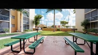 This outdoor common area is a welcoming, community-focused space set against the backdrop of a multi-story residential building with warm ochre-toned facades and open balconies. The grounds are thoughtfully landscaped, featuring well-maintained green lawns interspersed with towering palm trees that lend a distinctly tropical character to the environment. Mulched planting beds frame the base of the palms, adding a tidy, finished look to the landscaping.

A spacious paved patio area serves as the centerpiece of the outdoor gathering space, furnished with multiple green metal mesh picnic tables and benches, providing ample seating for residents to relax and socialize. The terracotta-toned concrete surface of the patio complements the warm color palette of the surrounding building exterior.

A dedicated single-story amenity building is situated on the grounds, constructed of red brick with a classic red clay tile roof. Large glass-front doors and windows reveal fitness equipment inside, identifying it as a resident fitness center. A paved walkway with decorative brick accents leads directly to its entrance, flanked by neatly trimmed hedges and potted greenery.

Paved pathways wind throughout the grounds, connecting the various outdoor areas and providing easy pedestrian access. The open sky above and surrounding vegetation create a bright, airy atmosphere that enhances the overall appeal of this community outdoor space.
