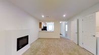 This single-floor space presents a bright and functional layout centered around a spacious living area finished with wall-to-wall carpeting in a warm beige tone. The room is anchored by a sleek, built-in fireplace along one wall, offering a focal point that adds both visual interest and comfort to the space. Recessed lighting in the smooth white ceiling provides even illumination throughout, contributing to the clean and airy atmosphere.

A pass-through window connects the living area to the kitchen, which features medium-toned wood cabinetry and a window that draws in natural light. This open-style connection between the two areas promotes a sense of openness and ease of interaction between spaces. The kitchen appears to include standard appliances and a sink positioned beneath the cabinetry.

A hallway visible from the living area leads to additional rooms, each benefiting from natural light filtering through windows. A closet with a white paneled door is also accessible from the main corridor. Throughout, the space maintains a consistent neutral color scheme with white walls and light trim, creating a versatile and move-in-ready environment.