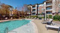 This outdoor pool area in Columbia, SC offers a resort-style retreat designed for relaxation and leisure. At its center sits a generously sized freeform swimming pool with crystal-clear blue water, accessible via stainless steel entry rails and featuring clearly marked depth gradients along its tiled border. The surrounding concrete sun deck provides ample space for sunbathing and socializing, furnished with multiple chaise lounges and small accent tables positioned throughout the area.

A dedicated outdoor dining and lounge zone is equipped with slatted patio tables, chairs, and a large shade umbrella, offering a comfortable spot for gathering. The pool area is bordered by meticulously maintained landscaping, including manicured shrubs, ornamental grasses, seasonal flowering plants, and mature trees that provide natural shade and privacy. Stone retaining walls with tiered plantings add architectural character to the perimeter.

Adjacent to the pool is a brick-clad community building with large windows, likely serving as a clubhouse or amenity facility. Classic-style lamp posts contribute to the refined aesthetic of the space. The entire area is enclosed by wrought iron fencing, ensuring security while maintaining open sightlines. Tall pine trees frame the backdrop, and the abundant natural sunlight throughout creates a bright, welcoming atmosphere year-round.