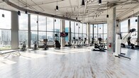 Located at 641 Division Street in Nashville's vibrant Gulch neighborhood, this expansive fitness center delivers a premium workout experience within a striking modern setting. The facility is defined by soaring ceilings and dramatic floor-to-ceiling glass curtain walls that wrap around multiple zones, flooding the interior with abundant natural light and offering sweeping panoramic views of the Nashville skyline.

The cardio area is a standout feature, equipped with a comprehensive lineup of treadmills, ellipticals, stationary bikes, stair climbers, and rowing machines, all positioned to take full advantage of the cityscape beyond the glass. Light hardwood-style flooring throughout the main floor lends a clean, airy aesthetic that complements the industrial-modern architecture, including exposed ductwork and concrete structural columns.

A dedicated strength and functional training zone features cable machines, free-motion equipment, a full dumbbell rack, kettlebells, medicine balls, and a power rack. A separate room with rubberized flooring is designed for high-intensity training, housing a heavy punching bag, BOSU balls, and additional free weights. Wall-length mirrors enhance the sense of space in this area.

The facility also includes a bright entry vestibule with curved glass walls and access to an outdoor terrace. Playful signage and thoughtful design details contribute to an energetic, welcoming atmosphere throughout.