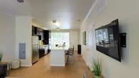 This single-floor residence showcases an open-concept layout that seamlessly connects the kitchen and living areas, creating a spacious and functional environment. The kitchen is outfitted with dark espresso cabinetry that provides a striking contrast against the light-colored walls and warm hardwood-style flooring running throughout. Stainless steel appliances, including a full-sized refrigerator, complement the modern aesthetic, while light countertops add brightness to the workspace.

A central kitchen island with a white finish serves as the focal point of the space, offering additional prep area and seating with contemporary bar-height chairs. This island also acts as a natural divider between the kitchen and the adjoining living area, maintaining an open and airy feel. Crown molding along the ceiling adds a refined architectural touch throughout.

Natural light flows generously into the space through large windows fitted with horizontal blinds, offering views of the surrounding greenery. A wall-mounted flat-screen television is positioned on the right side of the room, further defining the living area. Decorative accents, including framed artwork and potted plants, contribute to a clean and welcoming ambiance overall.