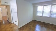 This single-floor space showcases a straightforward and functional layout designed to maximize usability. The living area is characterized by warm, classic parquet wood flooring arranged in a distinctive block pattern, adding a timeless aesthetic to the space. Neutral white walls throughout create a clean, bright backdrop that offers versatility for personalization. A window fitted with horizontal blinds allows natural light to filter into the living area, providing a connection to the outdoors while maintaining privacy. The open passageway connects the main living space seamlessly to the kitchen area, which features white cabinetry and a full-size refrigerator, contributing to the unit's cohesive, uncluttered feel. White baseboards frame the perimeter of the rooms, adding a crisp finishing detail that complements the overall neutral palette. The layout is compact yet practical, making efficient use of the available space. The consistent flooring and color scheme across visible areas lend the space a unified, easy-to-maintain character suitable for everyday living.