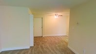 This single-floor space presents a practical and straightforward layout centered around a modest main living area. The room is finished with wall-to-wall beige carpeting that extends throughout, providing a warm and cohesive feel underfoot. Neutral, light yellow-toned walls reflect ambient light and lend the space a simple, understated aesthetic that serves as a blank canvas for personalization.

A ceiling fan with an integrated light fixture is centrally mounted, offering both overhead illumination and air circulation. White baseboards run along the perimeter of the room, adding a clean contrast to the carpet and walls. The entry door is positioned along one wall and is framed by a partial wall partition, subtly defining the entryway from the main living space.

The layout is open and unobstructed, allowing for flexible furniture arrangement. The overall ambiance is functional and understated, suited for those seeking a low-maintenance, move-in-ready living environment. The space reflects a classic, no-frills design that prioritizes practicality while maintaining a clean and livable atmosphere.
