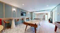 Located at 808 Chapel Street in the heart of New Haven, CT, this thoughtfully designed amenity floor offers a generously sized, single-level space built for socializing and relaxation. The interiors are defined by a cohesive modern aesthetic, with soft teal and sage green accent walls, geometric patterned tile flooring, and warm wood-tone finishes throughout.

The centerpiece of the space is a lively game lounge featuring an air hockey table, a wall-mounted flat-screen TV, a built-in red banquette, and high-top bar seating with warm wall sconces — all framed by elegant paneled wainscoting. Adjacent to the lounge is a fully equipped communal kitchen and dining area, complete with a long island bar with seating, a textured tile backsplash, stainless steel appliances, and a retro-style refrigerator. Yale-themed décor adds a nod to the local collegiate culture.

The hallways connect to an inviting lobby entry featuring bold geometric tile floors and a dramatic floor-to-ceiling living green wall, accented by vibrant orange pod chairs that create a striking visual focal point.

An outdoor terrace rounds out the amenities, offering a warm-weather retreat with wood deck flooring, lush planters, colorful lounge seating, and dining tables set against an urban brick backdrop.