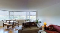 This single-floor space is defined by its striking curved corner window arrangement, which dominates the main living area and floods the interior with natural light. The panoramic windows frame lush, verdant treetop views, creating a strong visual connection to the outdoors and lending the space a serene, elevated ambiance.

The living area is anchored by a neutral-toned sofa accented with decorative throw pillows and a richly patterned textile blanket, complemented by warm accent lighting from a table lamp on a wooden side table. A tall indoor palm adds a natural, organic touch to the corner of the room, enhancing the connection to the greenery visible through the windows.

Adjacent to the living area, a compact dining nook accommodates a round table with wooden chairs, positioned to take full advantage of the corner window views. The flooring throughout has a warm, polished tone that reflects the abundant natural light.

The walls are finished in clean white, contributing to an open, airy feel throughout the space. Roller shades on the windows provide privacy and light control while maintaining the room's minimalist aesthetic. Overall, the space offers a comfortable and light-filled environment with a notable emphasis on natural scenery.