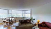 This single-floor space is defined by its striking curved corner window arrangement, which dominates the main living area and floods the interior with natural light. The panoramic windows frame lush, verdant treetop views, creating a strong visual connection to the outdoors and lending the space a serene, elevated ambiance.

The living area is anchored by a neutral-toned sofa accented with decorative throw pillows and a richly patterned textile blanket, complemented by warm accent lighting from a table lamp on a wooden side table. A tall indoor palm adds a natural, organic touch to the corner of the room, enhancing the connection to the greenery visible through the windows.

Adjacent to the living area, a compact dining nook accommodates a round table with wooden chairs, positioned to take full advantage of the corner window views. The flooring throughout has a warm, polished tone that reflects the abundant natural light.

The walls are finished in clean white, contributing to an open, airy feel throughout the space. Roller shades on the windows provide privacy and light control while maintaining the room's minimalist aesthetic. Overall, the space offers a comfortable and light-filled environment with a notable emphasis on natural scenery.
