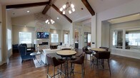 This single-story gathering space combines rustic and contemporary design elements to create an inviting, open atmosphere. The room's most striking feature is its soaring vaulted ceiling, accented by rich, dark-stained exposed wood beams that draw the eye upward and add warmth to the space. Two stylish chandeliers with clustered globe pendants hang from the ceiling, providing both ambient and focused lighting throughout the room.

The focal point of the lounge area is a floor-to-ceiling stacked stone fireplace flanked by large windows that allow ample natural light to fill the space. A wall-mounted television above the fireplace adds a functional entertainment element. The hardwood flooring throughout reinforces the warm, welcoming aesthetic.

Seating is thoughtfully arranged to accommodate various uses. Comfortable upholstered sofas and chairs create a relaxed conversation area near the fireplace, while bistro-style high-top tables paired with leather-upholstered chairs offer casual seating options toward the center of the room. A sliding barn door in the background hints at additional spaces beyond, adding an architectural detail that complements the overall rustic-modern design theme.
