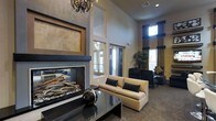 This single-floor space exudes sophistication through its thoughtful design and upscale finishes. The centerpiece of the main lounge area is a striking floor-to-ceiling fireplace framed in sleek dark stone and accented by warm travertine tile, providing both visual drama and a cozy focal point. Soaring ceilings amplify the sense of openness, while a decorative crystal chandelier adds a touch of elegance overhead. The seating area features a cream upholstered sofa with dark accent pillows, paired with contemporary armchairs to create an inviting conversational arrangement. A dark, reflective coffee table anchors the space and complements the overall neutral-toned palette of warm beiges and soft grays. Natural light filters in through tall windows dressed in rich navy blue drapery, lending depth and color contrast to the room. Lush indoor greenery adds a refreshing organic element, softening the polished interior. The overall ambiance strikes a balance between modern refinement and comfortable warmth, making it a welcoming and functional gathering space suited for relaxation or socializing.