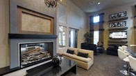 This single-floor space exudes sophistication through its thoughtful design and upscale finishes. The centerpiece of the main lounge area is a striking floor-to-ceiling fireplace framed in sleek dark stone and accented by warm travertine tile, providing both visual drama and a cozy focal point. Soaring ceilings amplify the sense of openness, while a decorative crystal chandelier adds a touch of elegance overhead. The seating area features a cream upholstered sofa with dark accent pillows, paired with contemporary armchairs to create an inviting conversational arrangement. A dark, reflective coffee table anchors the space and complements the overall neutral-toned palette of warm beiges and soft grays. Natural light filters in through tall windows dressed in rich navy blue drapery, lending depth and color contrast to the room. Lush indoor greenery adds a refreshing organic element, softening the polished interior. The overall ambiance strikes a balance between modern refinement and comfortable warmth, making it a welcoming and functional gathering space suited for relaxation or socializing.