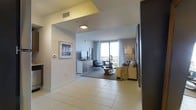 This single-floor space is defined by a clean, contemporary design that begins at the entryway, where large-format light-tiled flooring guides the eye toward the main living area. The transition from the entry hall into the living room is framed by a softly curved archway, adding architectural interest while maintaining an open, connected feel throughout.

The living area is generously proportioned and centers around a neutral-toned sofa ensemble paired with a round coffee table atop a dark area rug, creating a well-defined seating zone. A flat-screen TV is mounted atop a low-profile media dresser along one wall, complemented by framed artwork that adds a curated, residential touch. A thermostat panel near the entryway highlights the unit's practical, modern amenities.

Natural light is a standout feature, with floor-to-ceiling windows spanning the exterior wall and offering direct access to a private balcony with open views. Sheer curtains allow diffused light to fill the space throughout the day, enhancing its bright and airy atmosphere. The overall palette of whites, grays, and warm neutrals contributes to a cohesive, understated elegance throughout.