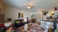 This single-floor space showcases an open-concept layout that seamlessly connects the living, dining, and kitchen areas, creating a cohesive and functional environment. The living area is anchored by a dark leather sofa accented with colorful throw pillows and a bold floral-patterned area rug, adding warmth and visual interest to the space. A woven drum stool serves as a decorative accent piece, and contemporary wall art lends a modern touch to the neutral, light-colored walls. Two ceiling fans with integrated lighting fixtures are positioned throughout the open space, ensuring comfortable airflow and ambient illumination. The dining area accommodates a dark wood table and matching chairs, providing a defined eating space within the open floor plan. Adjacent to the dining area, the kitchen features a raised bar counter with seating, offering additional dining options. Closet doors visible in the background suggest built-in storage solutions. The light tile flooring runs consistently throughout, unifying the space and contributing to its bright, airy feel. Overall, the layout is practical and well-organized, making efficient use of the available space.