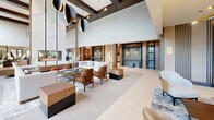 Located at 500 Parq Ln in Parsippany, NJ, this impressive single-floor common area showcases a thoughtfully designed, upscale residential lobby environment spanning a generous footprint. The space is defined by soaring double-height ceilings, exposed dark wood beam accents, and dramatic floor-to-ceiling windows that flood the interior with abundant natural light while offering views of the surrounding landscaped grounds.

The open-plan layout is organized into several distinct yet seamlessly connected zones. A welcoming reception area greets visitors with a sleek front desk framed by warm wood paneling and a striking marble-clad fireplace wall. Adjacent seating areas feature curated arrangements of modern lounge chairs in rich blue and earthy tan tones, set atop layered area rugs over large-format light stone tile flooring. Sculptural pendant light fixtures with brass and matte black finishes add an artistic dimension throughout.

A communal dining and social area is outfitted with a long bar-height table and seating for many, anchored by contemporary pendant lighting and flanked by views to an outdoor terrace. Decorative gold-framed mirrors, curated artwork, and accent furniture in walnut and brass tones elevate the overall aesthetic. Elevator bays with distinctive glass block surrounds add architectural interest. The overall ambiance is polished, contemporary, and inviting.