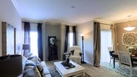 This single-floor space showcases a thoughtfully designed living room that balances comfort and style. The room is finished with classic crown molding along the ceiling perimeter, adding a refined architectural detail that elevates the overall aesthetic. Soft, neutral-toned walls create a calm and inviting atmosphere throughout.

The living area is carpeted in a warm, plush material that contributes to the cozy ambiance. Multiple windows along the walls allow generous amounts of natural light to fill the space, dressed with dark curtain panels that provide a striking contrast against the lighter walls and offer adjustable privacy. Vertical blinds on one window add a practical touch.

Furnishings include a comfortable sectional sofa, a wicker accent chair with a draped throw, and a white coffee table serving as a central focal point. A tall torchiere floor lamp provides additional warm ambient lighting. A decorative dark cabinet with a framed mirror rests against one wall, adding character and depth to the room. The space is accessorized with carefully placed décor, reflecting an attentive and curated interior style.