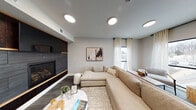 This contemporary single-floor space is thoughtfully designed to balance comfort, style, and functionality. The centerpiece of the main lounge area is a bold, dark slate-tiled fireplace wall accented with warm wood shelving and cabinetry, creating a striking focal point that anchors the room. A large, plush sectional sofa in neutral tones is positioned around a round marble-top coffee table atop a textured area rug, offering an inviting and relaxed gathering environment.

Opposite the fireplace wall, an open industrial-style shelving unit crafted from natural wood and black metal provides generous display and storage space, adding an earthy, organic contrast to the room's modern palette. The flooring throughout features patterned carpet in cool grey tones, complementing the soft grey walls and clean white ceiling fitted with recessed circular light fixtures.

Toward the windowed end of the space, a seating nook with modern accent chairs and a built-in counter-height workstation offers a versatile area for both relaxed conversation and focused work. A generous bank of large windows dressed with sheer cream curtains floods this zone with natural light and frames views of the surrounding neighborhood. A glass-paneled interior door adds a refined architectural touch, enhancing the open, airy ambiance throughout the space.