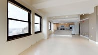 This single-floor space offers an open-plan layout that seamlessly connects the living area to a well-appointed kitchen. Large, black-framed windows flood the interior with natural light while framing striking elevated city views, including glimpses of surrounding urban architecture. The pale tile flooring runs continuously throughout, reinforcing a sense of spaciousness and visual cohesion across the entire floor plan.

The kitchen is positioned toward the rear of the space and features light wood cabinetry, a dark countertop island with a breakfast bar overhang, and stainless steel appliances including a refrigerator and microwave. Overhead pendant lighting and recessed track lighting provide practical illumination in the cooking area.

The ceiling is defined by structural beams finished in white, adding architectural character while maintaining a clean, contemporary aesthetic. The neutral white walls and light flooring create a bright, airy ambiance throughout. The open layout, elevated position, and abundance of natural light make this a versatile and visually appealing urban space.