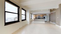 This single-floor space offers an open-plan layout that seamlessly connects the living area to a well-appointed kitchen. Large, black-framed windows flood the interior with natural light while framing striking elevated city views, including glimpses of surrounding urban architecture. The pale tile flooring runs continuously throughout, reinforcing a sense of spaciousness and visual cohesion across the entire floor plan.

The kitchen is positioned toward the rear of the space and features light wood cabinetry, a dark countertop island with a breakfast bar overhang, and stainless steel appliances including a refrigerator and microwave. Overhead pendant lighting and recessed track lighting provide practical illumination in the cooking area.

The ceiling is defined by structural beams finished in white, adding architectural character while maintaining a clean, contemporary aesthetic. The neutral white walls and light flooring create a bright, airy ambiance throughout. The open layout, elevated position, and abundance of natural light make this a versatile and visually appealing urban space.