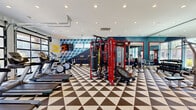 Located in Hammond, Louisiana, this fitness center offers a generously sized, single-floor exercise room designed to support a full range of workout routines. The space makes a strong visual impression with its bold, geometric-patterned flooring in contrasting black and white triangles, complemented by deep navy and teal accent walls adorned with a striking three-dimensional cube mural. Recessed ceiling lighting throughout ensures the space is evenly and brightly lit, while motivational neon signage adds an energetic touch to the atmosphere.

Two large glass garage-style doors line one wall, flooding the room with natural light and offering open views of the surrounding outdoor environment — creating an airy, inviting workout setting. The cardio zone features multiple treadmills and elliptical machines positioned to take advantage of these views. A dedicated cycling area includes spin bikes alongside strength machines and a wall-mounted flat-screen TV.

The strength training section is anchored by a prominent red multi-functional cable machine system, accompanied by a heavy punching bag, free weights, a cable row machine, and a power rack. Floor-to-ceiling mirrors along one wall visually expand the space and allow users to monitor their form. The combination of modern equipment, dynamic design, and ample natural light makes this fitness center a vibrant and functional training environment.