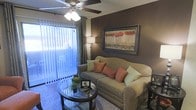 This single-floor residential space offers a comfortable and thoughtfully designed living environment. The living room serves as the heart of the home, showcasing a warm and inviting ambiance through its rich, earthy color palette. A deep brown accent wall provides a striking backdrop for a framed piece of wall art featuring bold red and neutral tones, adding a pop of color and artistic character to the space. The room is furnished with a plush, neutral-toned sofa adorned with warm orange and green accent pillows, creating a relaxed yet stylish seating area. A round glass-top coffee table sits at the center, decorated with small greenery and personal accents that add a homey touch. Soft carpeting underfoot enhances the cozy feel of the room. Natural light pours in abundantly through a large sliding glass door dressed with vertical blinds and elegant drape panels, offering a bright and airy atmosphere. A ceiling fan with an integrated light fixture is centrally positioned, ensuring both comfort and practical illumination. A tall floor lamp in the corner provides additional warm ambient lighting, rounding out the well-layered lighting scheme throughout the living area.