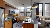 This thoughtfully designed single-floor residence showcases a contemporary aesthetic throughout, blending warm wood tones with sleek finishes and an abundance of natural light. The kitchen is a standout feature, equipped with floor-to-ceiling warm wood cabinetry, dark stone countertops, full stainless steel appliances including a refrigerator, electric range, and over-the-range microwave, and a peninsula with pendant lighting overhead. Black-framed windows allow natural light to pour in, offering views of the surrounding neighborhood.

The living and dining area is distinguished by dramatically tall black-framed windows draped with floor-length curtains, creating a striking sense of height and openness. Natural light floods this space, enhancing its airy, urban character. A separate dining nook accommodates intimate gatherings, while a dedicated workspace features a rustic wood table complemented by a large triptych landscape artwork.

The bedroom is comfortably proportioned, finished with plush carpeting and clean white walls. The bathroom features dark tile flooring, a frameless glass walk-in shower with contrasting light subway tile, and modern fixtures. A hallway houses a convenient stacked in-unit washer and dryer, maximizing space efficiency throughout.