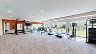 Located at 601 Industrial Blvd W in Waconia, Minnesota, this fitness center offers a comprehensive workout facility spread across a single, generously sized floor. The space is divided into two distinct areas: a main exercise room and a flexible studio room, each thoughtfully designed to support a wide range of fitness activities.

The main exercise room is bright and open, featuring large windows along one wall that flood the space with natural light and provide views of the surrounding greenery. The room is outfitted with a full array of cardio equipment, including treadmills, ellipticals, rowing machines, and a stationary bike, as well as strength training options such as a power rack, cable machine, TRX suspension system, kettlebells, medicine balls, and a dumbbell rack. A bold orange accent wall adds energy to the space, and a wall-mounted flat-screen TV enhances the workout experience. Patterned carpet flooring, recessed lighting, and a water fountain station contribute to the room's functional and welcoming atmosphere.

The adjoining studio room offers a serene environment for yoga, stretching, or group classes. It features light hardwood-style flooring, floor-to-ceiling mirrors, and expansive windows overlooking lush outdoor scenery. A sliding barn door connects the two spaces seamlessly, maintaining an open and versatile flow throughout the facility.