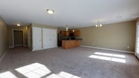 Located at 526 Middletown Rd in Hummelstown, PA, this single-floor unit offers an open-concept layout that seamlessly connects the kitchen and living areas into one expansive, functional space. The interior is finished with neutral beige walls and white trim, creating a clean and welcoming ambiance throughout.

The kitchen is positioned centrally within the open floor plan and features warm honey-toned wood cabinetry, dark countertops, and a peninsula with additional counter seating and storage. Black appliances, including a refrigerator and microwave, are integrated into the cabinetry. Pendant lighting over the kitchen area and a chandelier in the adjoining living space provide layered illumination, complementing the natural light that filters in from windows along the far wall.

The carpeted living area is generously sized, offering flexibility for various furniture arrangements. Along one wall, two large closets with paneled white doors provide substantial built-in storage. The space maintains a simple, practical aesthetic with consistent flooring and ceiling height throughout, making it well-suited for everyday living.