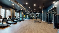 Located at 200 E Cama St in Charlotte, NC, this single-floor fitness center offers a comprehensive and thoughtfully designed workout facility. The space is defined by its striking teal-blue walls, dark ceilings with exposed industrial ductwork, and warm wood-look flooring — a combination that gives the gym a modern yet inviting atmosphere. Recessed and accent lighting throughout creates a focused, energized environment.

The cardio zone features a row of treadmills and elliptical machines, positioned alongside large windows dressed with linen curtains that allow natural light to fill the space. A dedicated strength training area is outfitted with Matrix cable machines, a multi-functional weight bench, and additional resistance equipment, all reflected in large, white-framed wall mirrors that amplify the sense of space. Medicine balls and stability equipment add versatility for functional fitness training.

A built-in wooden shelving and cabinetry unit provides organized storage for accessories and equipment. Decorative wall sconces and a colorful abstract painting contribute to the polished aesthetic. Black-framed glass double doors open to an adjacent lounge area, seamlessly connecting the fitness center to the broader facility. Ceiling fans and ample ventilation ensure comfort during workouts. Overall, the space balances high functionality with refined design.