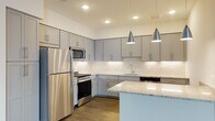 This well-appointed single-floor residence showcases a thoughtfully designed open-concept layout that seamlessly connects the kitchen and living areas. The kitchen is a standout feature, outfitted with soft gray shaker-style cabinetry, granite countertops, a classic white subway tile backsplash, and a full suite of stainless steel appliances including a gas range, built-in microwave, and refrigerator. Stylish pendant lighting hangs above the kitchen island, adding both function and ambiance. Rich, dark hardwood-style flooring flows throughout the main living areas, complementing the neutral color palette and creating a warm, cohesive aesthetic. Natural light enters through a large window in the spacious living area, enhancing its open and airy feel. The primary bedroom features plush carpeting and direct access to a generously sized walk-in closet with multi-level wire shelving. Two bathrooms serve the residence: one with a subway-tiled soaking tub and shower combination, and another featuring a standalone glass-enclosed shower with light tile flooring. Granite vanity countertops and gray cabinetry carry the kitchen's design language throughout. A dedicated in-unit laundry area with a stacked washer and dryer adds everyday convenience to this modern, move-in-ready space.