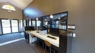This single-story community amenity space features a thoughtfully designed internet café area that blends functionality with contemporary style. The internet café is anchored by a clean, white countertop workstation lined with desktop computers, a printer, and cushioned bar-height stools, providing residents with a convenient workspace. Pendant globe lights hang overhead, casting warm, focused illumination across the counter and enhancing the modern aesthetic.

Dark cabinetry and wall paneling contrast sharply with the bright countertop, creating a polished, professional atmosphere. Large glass windows along one wall offer a clear view into an adjacent fitness center equipped with Precor cardio machines, connecting the two amenity spaces visually while maintaining separation. Natural light filters in through an exterior entrance nearby, brightening the space and adding openness.

The flooring transitions between tile and carpet, subtly defining different zones within the area. Vaulted ceilings add a sense of vertical spaciousness to the room. Overall, the space presents a clean, well-appointed amenity hub designed to support both productivity and wellness.