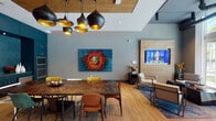 This sophisticated single-floor amenity space seamlessly blends modern design with a warm, welcoming ambiance. The open-plan layout connects a spacious lounge area, a dining zone, and a fully equipped kitchen, all unified by wide-plank hardwood flooring and soaring ceilings that lend a generous sense of volume throughout.

The lounge area is anchored by a wall-mounted flat-screen TV set into a warm wood-paneled accent wall, surrounded by a curated arrangement of upholstered armchairs and sculptural geometric coffee tables atop richly toned area rugs. Expansive floor-to-ceiling glass doors flood the space with natural light and open directly onto an outdoor terrace, offering views of palm trees and the surrounding urban skyline.

The dining area features a striking live-edge wood table paired with an eclectic mix of colorful mid-century chairs, all beneath a dramatic cluster of matte black and gold pendant lights. A partial wood-panel ceiling detail above adds warmth and visual depth to this zone.

The kitchen is a standout feature, finished in bold deep teal cabinetry with a textured geometric tile backsplash. Built-in appliances, ample counter space, and a full sink make it well-suited for entertaining and events.

Throughout the space, bold original artworks add personality and vibrancy, while the industrial-style exposed ceiling elements in the lounge contribute an urban, contemporary edge. A black-framed glass partition wall leads to an adjacent corridor, maintaining visual openness while defining distinct functional zones.