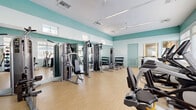 Located at 8485 Hammocks Boulevard in Miami, FL, this fitness center offers a well-organized and comprehensively equipped workout facility spread across a single floor. The space is defined by its bright, airy atmosphere, enhanced by soft aqua-teal accent walls that contrast with crisp white ceilings and light hardwood-style flooring throughout.

The main gym area is generously outfitted with a wide range of equipment to support diverse workout routines. Strength training is well-represented with multiple cable and weight machines, a full dumbbell rack, and a flat bench, while large wall-mounted mirrors amplify the sense of space and allow for proper form checks. The cardio section features a row of elliptical trainers and treadmills positioned along the windowed wall, benefiting from ample natural light streaming through multiple windows fitted with slatted blinds.

A separate room houses a dedicated cycling studio with spin bikes, providing a more focused environment for indoor cycling sessions. Additional accessories such as stability balls and resistance equipment are also available throughout the space.

Ceiling fans and a modern linear lighting system ensure the environment remains comfortable and well-lit. The overall design is clean, functional, and welcoming, making this fitness center a practical and inviting resource for residents in the Hammocks community of Miami.