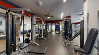 Located at 322 Baldwin Rd in Odenton, Maryland, this well-appointed fitness center offers a generous single-floor layout designed to accommodate a comprehensive range of workout needs. The space is thoughtfully divided into distinct strength and cardio zones, providing a functional and organized environment for users of all fitness levels.

The strength training area is outfitted with a robust selection of cable machines, multi-functional cable crossover systems, free weights, adjustable benches, and weight racks. Large wall-mounted mirrors amplify the sense of space and allow users to monitor their form throughout their workouts. Bold red and dark gray accent bands run along the upper walls, giving the space a dynamic, energetic aesthetic.

The cardio zone features treadmills, elliptical machines, rowing machines, and stationary spin bikes equipped with individual screens. This section benefits from floor-to-ceiling glass windows and sliding glass doors that flood the area with natural light and offer views of the surrounding greenery, creating an open and inviting atmosphere.

Throughout the facility, durable rubber and hardwood-style flooring is used to define different activity areas. Ceiling fans and recessed linear lighting ensure proper ventilation and visibility. A wall-mounted television is also present for entertainment during workouts. The overall design balances functionality with a modern, motivating ambiance.