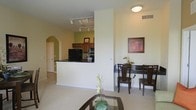 This single-floor residence showcases an open-concept layout that seamlessly connects the main living areas. The kitchen is equipped with dark appliances, including a black refrigerator and range, complemented by warm wood cabinetry and a dark-topped breakfast bar that serves as a natural dividing point between the kitchen and the rest of the living space. An arched entryway adds architectural character, lending a soft, classic touch to the otherwise contemporary design.

The dining area sits adjacent to the main living space and comfortably accommodates a dark wood dining table and chairs. Decorative wall art and a small plant centerpiece on the dining table add warmth and personality to the space. Neutral, light-toned carpet flows throughout the living and dining areas, contributing to a cohesive and inviting ambiance.

The living area, partially visible in the foreground, features a glass-topped coffee table accented with floral décor. Recessed track lighting on the ceiling provides adjustable illumination throughout, while a flush-mount ceiling fixture in the dining area ensures even lighting. The overall color palette of soft beiges and greens creates a calm, welcoming atmosphere suited for everyday living.