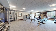 This fitness center is a comprehensively appointed facility designed to support a wide range of workout routines. The main exercise floor is generously proportioned, featuring patterned carpet flooring, a drop tile ceiling with recessed lighting, and large windows that invite natural light and outdoor views. The room houses an impressive array of equipment, including cable machines, selectorized strength machines, ellipticals, stationary bikes, treadmills, and adjustable benches, along with a dedicated dumbbell rack set against a bold blue accent wall with a wall-mounted television.

A separate studio room offers a clean, open space finished with wood-look flooring, floor-to-ceiling mirrors with a barre rail, a wall-mounted TV, and an abundance of accessories including resistance bands, stability balls, foam rollers, and yoga mats — ideal for group fitness, stretching, or personal training sessions.

The facility also features an elegantly appointed entry corridor with rich dark wood wainscoting and framed artwork, lending a refined aesthetic to the space. A water station with a dual drinking fountain and bottle filler, set against a striking dark tile brick wall, adds a practical and visually distinctive touch. Throughout the space, sanitizing stations and thoughtful detailing reflect a commitment to cleanliness and user comfort.