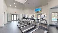 Located at 675 Jamestown Blvd in Altamonte Springs, FL, this fitness facility offers a comprehensive, single-floor layout designed to accommodate a wide range of exercise needs. The main exercise room impresses with soaring vaulted ceilings and an abundance of natural light streaming through large windows and glass double doors. The open floor plan is outfitted with a full lineup of TRUE-brand cardio equipment, including treadmills, stationary bikes, and elliptical machines, complemented by a cable weight machine, free weights, and kettlebells. Durable rubber sport flooring spans the cardio and strength areas, while full-length mirrors enhance the sense of space. A dedicated studio room features wood-look flooring, floor-to-ceiling mirrored walls, tray ceilings with teal crown molding, and multiple windows offering views of the surrounding greenery — ideal for yoga, stretching, or group fitness. This area also stores medicine balls, stability balls, and resistance equipment. A connecting hallway with wood-look flooring links the spaces and leads to a welcoming lounge area, complete with a ceiling fan, wall-mounted television, and a window with outdoor views. Together, these spaces create a bright, functional, and well-appointed fitness environment.