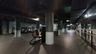 This fitness facility presents a striking industrial aesthetic defined by dark-painted exposed ceilings with visible ductwork and piping, structural columns finished in a two-tone grey and white treatment, and durable rubber flooring throughout. The space is generously proportioned, with the open floor plan allowing equipment zones to flow seamlessly into one another beneath consistent overhead lighting that punctuates the darker ambiance with focused illumination.

The cardio zone features a row of treadmills and a selection of elliptical trainers and stationary bikes, including rowing machines, all positioned to allow ample circulation space. The strength training area is well-equipped with cable machines, resistance equipment, power racks, and adjustable benches, complemented by a mirrored wall section that enhances the sense of depth. A dedicated boxing and functional training zone features heavy bags, open mat flooring, and wall-mounted equipment storage, accented by a distinctive botanical-patterned feature wall that adds a visual contrast to the otherwise utilitarian environment. A separate enclosed room with glass partition walls houses a refreshment station with a beverage vending machine and seating. The overall atmosphere is focused and purposeful, with a modern, no-frills design suited to serious training.
