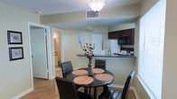 This single-floor residence offers a practical and comfortable layout designed for everyday living. The open-concept dining area flows seamlessly into the kitchen, creating a connected and functional living space. The dining area features warm hardwood flooring and is furnished with a round black dining table surrounded by matching black leather chairs, centered beneath a classic ceiling light fixture. A decorative floral arrangement adds a touch of warmth to the space.

The kitchen is thoughtfully designed with dark espresso-toned upper cabinetry and a dark granite-style countertop that extends into a breakfast bar, offering additional seating and serving space. The kitchen is equipped with essential appliances including a white refrigerator, an electric range, and a built-in microwave. A single-basin sink with a modern faucet is positioned at the bar counter for added convenience.

The textured ceiling and neutral wall tones throughout the space provide a clean and understated backdrop. A hallway visible from the dining area leads to additional rooms, including a bathroom, suggesting a well-organized floor plan. The overall ambiance is practical and inviting, with a layout that supports both daily routines and casual entertaining.