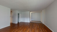 This single-floor unit offers a thoughtfully laid out living space defined by warm, dark hardwood flooring that extends throughout the main living areas. The expansive open-plan living room is a standout feature, showcasing a distinctive sunken carpeted section that creates a natural separation of space, adding architectural interest and a sense of depth. A large window allows natural light to filter into the living area, brightening the neutral gray walls and enhancing the overall sense of openness. A built-in closet with bifold doors provides convenient storage within the main living space.

The kitchen is efficiently designed, featuring warm honey-toned wood cabinetry with ample upper and lower storage, dark granite countertops, and a tile mosaic backsplash accent. Black appliances, including a gas range and a full-size refrigerator, complement the cabinetry, while light ceramic tile flooring contrasts neatly with the rest of the unit. A pass-through opening connects the kitchen to the living area, promoting an open feel.

The bathroom is finished with light ceramic tile flooring and wall tile detailing, including a decorative mosaic border accent. A rich wood vanity with a stone countertop and a chrome towel bar complete the space, lending a polished, cohesive look throughout.