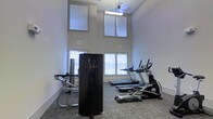 This fitness center is designed to provide a functional and comfortable workout environment. The space is immediately distinguished by its impressive double-height ceilings, which create an open, airy atmosphere and contribute to a sense of spaciousness. Large windows on multiple walls, fitted with blinds, allow generous amounts of natural light to fill the room while offering the flexibility to control glare during workouts. Overhead, a ceiling fan ensures proper air circulation throughout the space.

The fitness center is outfitted with a solid selection of exercise equipment. Cardio options include multiple treadmills and at least one elliptical machine, all positioned to face the windowed walls. Strength and resistance training is supported by a multi-function cable machine and additional equipment along the perimeter. A tall, freestanding storage or equipment unit is centrally positioned in the space.

The flooring consists of durable, textured carpet tile in a neutral tone, well-suited for a high-traffic fitness environment. The walls are finished in a clean, light neutral tone that complements the bright, motivating atmosphere. Overall, the fitness center offers a practical and well-lit setting suitable for a variety of workout routines.
