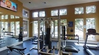 This fitness center offers a comprehensive workout environment designed to accommodate a wide range of exercise needs. The space is anchored by a multi-station cable machine and weight training equipment, complemented by a full dumbbell rack stocked with an extensive selection of free weights. Stability and medicine balls are also available, providing options for functional and core training.

The room features warm, earth-toned walls that create an energizing yet focused atmosphere. Large, multi-pane windows line the upper and lower portions of the walls on multiple sides, flooding the interior with abundant natural light and offering views of the surrounding greenery. The high ceiling height enhances the sense of openness and airflow throughout the space.

Dark rubberized flooring covers the workout area, providing both durability and safety underfoot. Wall-mounted mirrors are positioned strategically to allow users to monitor their form during exercises. Informational signage, including fitness center rules and guidelines, is posted near the entrance for easy reference.

Overall, this fitness center is a thoughtfully arranged, well-lit facility that provides members with the essential equipment and comfortable environment needed for an effective and well-rounded workout routine.