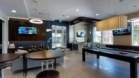 This well-appointed single-floor amenity space blends modern design with social functionality, making it an inviting destination for recreation and relaxation. The open-concept layout is anchored by a full-size pool table set atop light wood-style flooring, which flows seamlessly throughout the entire space. Bold navy blue accent walls contrast beautifully with the bright white ceilings, creating a dynamic and contemporary atmosphere. Recessed lighting, along with a striking circular pendant light and sleek hanging fixtures, ensures the space is warmly and evenly illuminated. Multiple wall-mounted flat-screen TVs are strategically positioned throughout the room, catering to both the lounge and game areas. A round dining table with modern bar-height seating occupies one corner, while a comfortable lounge area with upholstered chairs offers a more relaxed setting. A wet bar or kitchenette area with counter seating is tucked toward the rear of the space, providing a convenient spot for refreshments. Large windows and a glass-panel door invite abundant natural light and offer views of the outdoor surroundings, enhancing the open and airy feel of the interior. Decorative elements, including sculptural accents and greenery, add personality and warmth to this thoughtfully designed communal space.