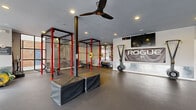 Located in Duluth, GA, this single-floor fitness center is a comprehensively outfitted exercise space designed for serious strength and conditioning training. The open layout is anchored by a series of Rogue power racks with red uprights, providing ample space for barbell work, including multiple loaded barbells with bumper plates ready for use. Rubber flooring spans the entire area, offering a durable and functional surface suited for heavy lifting and dynamic movements.

The space is well-lit with recessed ceiling lights supplemented by generous natural light streaming through large floor-to-ceiling windows and glass-panel doors, which open directly to an outdoor area. A ceiling fan keeps air circulating throughout. One wall features a mounted flat-screen TV alongside a prominent Rogue banner, adding an energetic visual focal point.

Equipment is thoughtfully organized throughout the space. A dedicated storage wall houses a full rack of kettlebells in varying weights alongside medicine balls and slam balls. Rogue plyo boxes are positioned centrally for plyometric training, and air assault bikes are available for cardiovascular conditioning. A cable/functional trainer and sled equipment round out the offerings. The space also benefits from access to an adjoining fitness area visible through interior glass doors, extending the overall training environment.